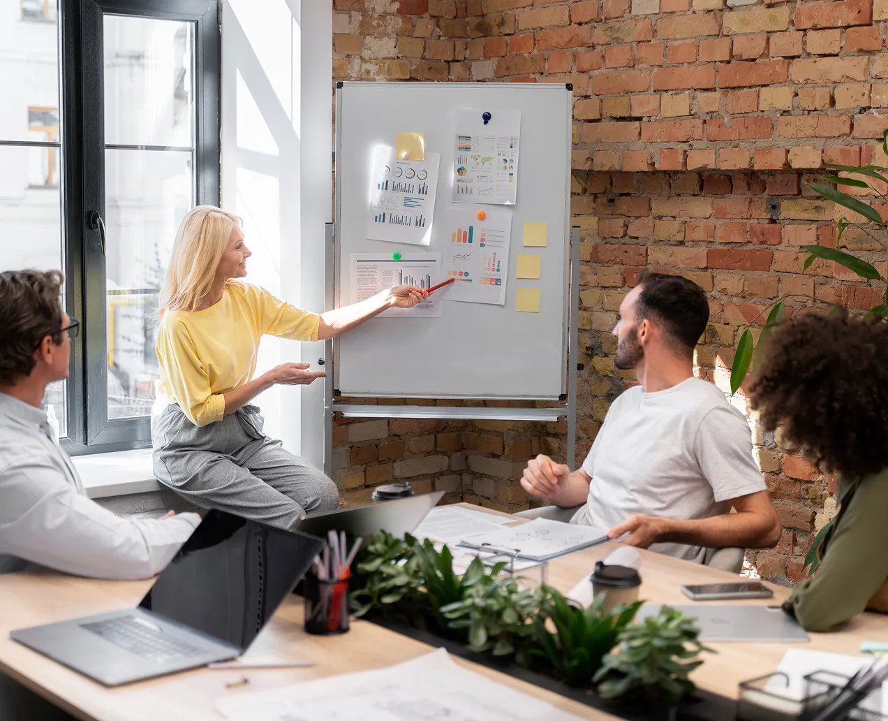A woman in a yellow shirt presenting charts on a whiteboard to three seated colleagues in a bright office with brick walls.